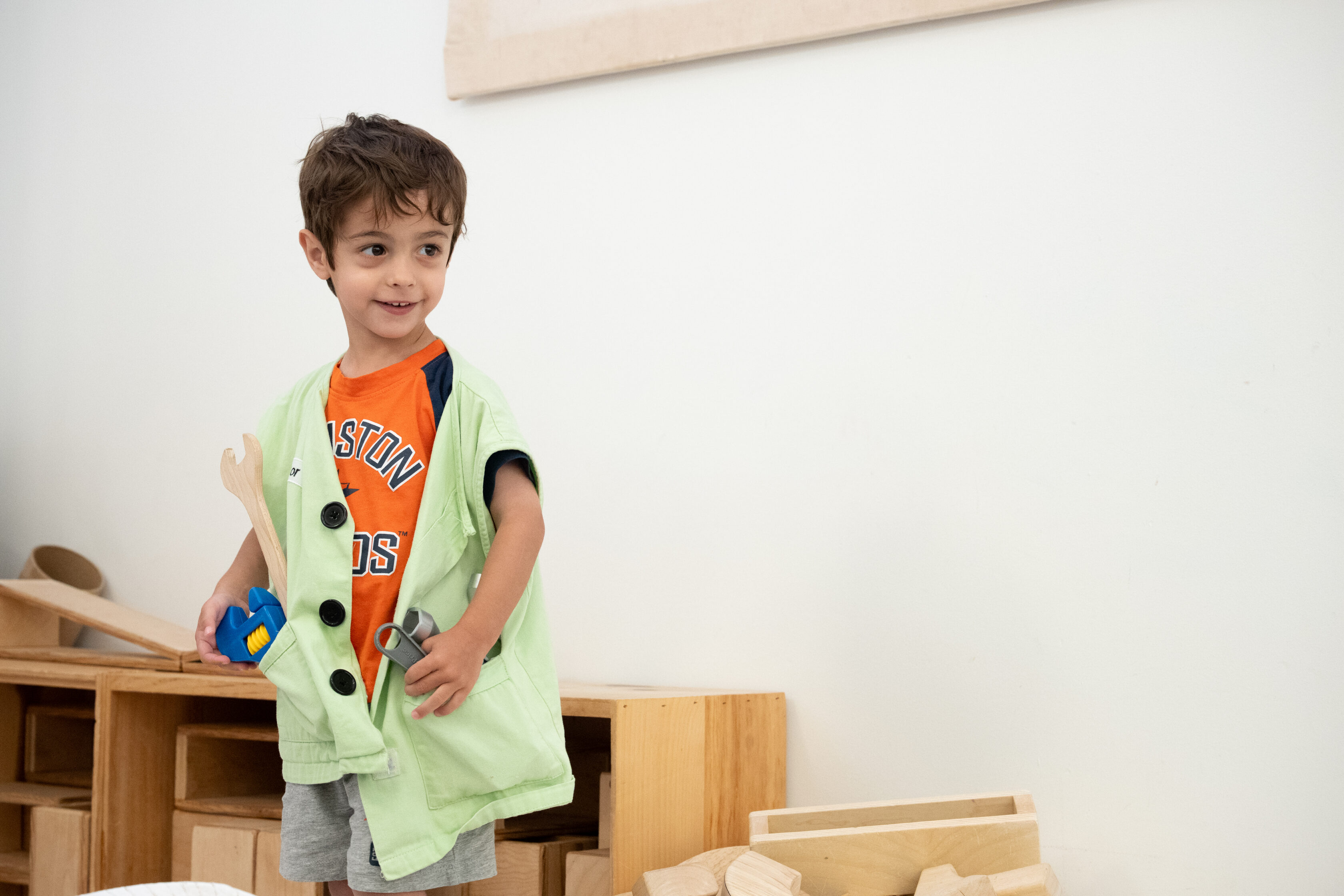 Young boy wearing a green vest and orange t-shirt stands in a play area holding wooden and plastic toys, with shelves of wooden blocks in the background.