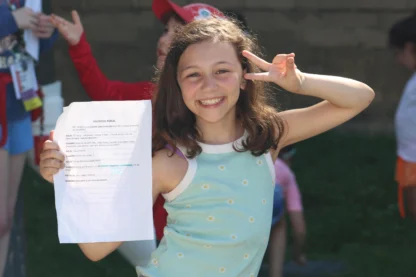 A girl smiles and holds up a piece of paper with writing on it, making a peace sign with her other hand, outdoors on a sunny day.