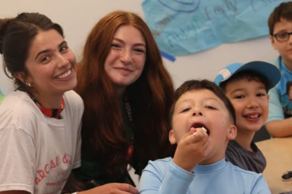 Four children sit at a table, two smiling at the camera, one eating, and one partially visible. Blue posters are hanging on the wall in the background.