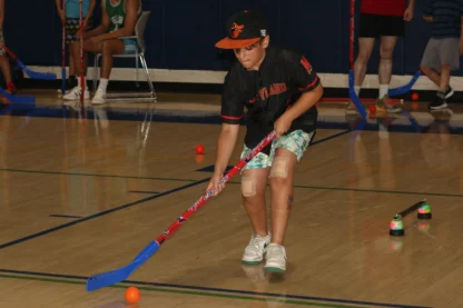 A boy wearing a black jersey and cap plays indoor floor hockey, focusing on the orange ball. Other players and hockey sticks are visible in the background.