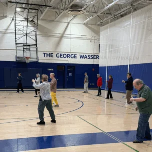 A group of people practice tai chi in a gymnasium, standing spaced apart on the basketball court under bright overhead lights.