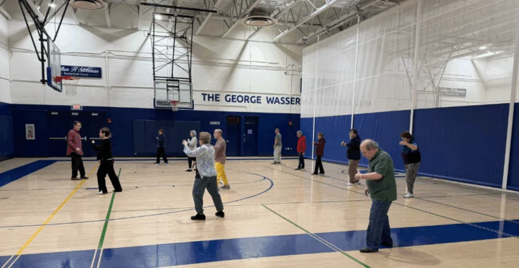 A group of people practice tai chi in a gymnasium, standing spaced apart on the basketball court under bright overhead lights.
