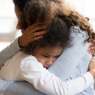 An adult and a child with curly hair sit on a couch, hugging closely. The child looks sad while the adult comforts them with an embracing hug.