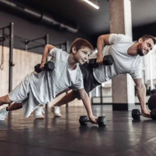 An adult and a child wearing white shirts do push-ups with dumbbells on a gym floor, surrounded by fitness equipment.