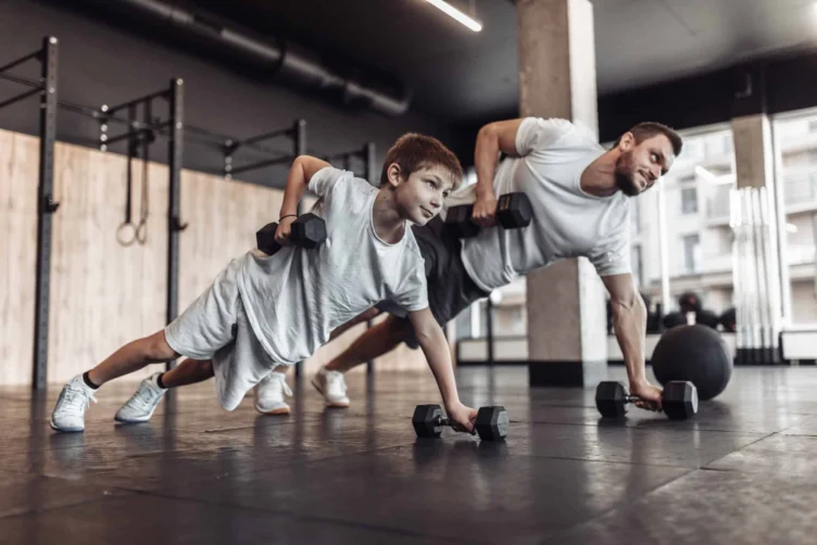 An adult and a child wearing white shirts do push-ups with dumbbells on a gym floor, surrounded by fitness equipment.