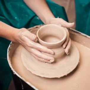 Hands shaping a clay cup on a pottery wheel, surrounded by wet clay and wearing a green apron.