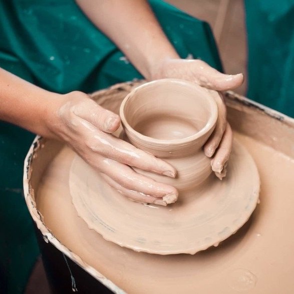 Hands shaping a clay cup on a pottery wheel, surrounded by wet clay and wearing a green apron.