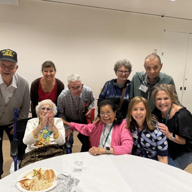 A group of older adults and caregivers pose and smile together around a table with food in a bright indoor setting.