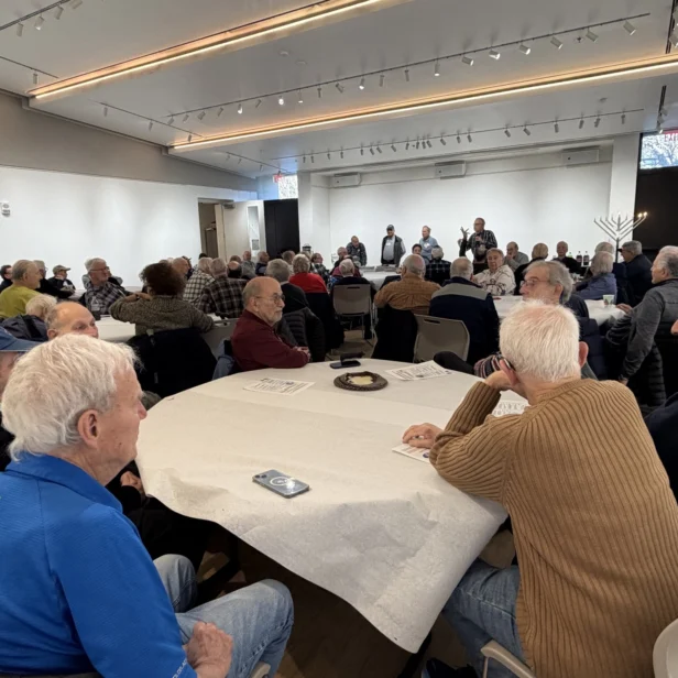 A group of older adults sit around tables in a large, well-lit room, attentively listening to speakers at the front.