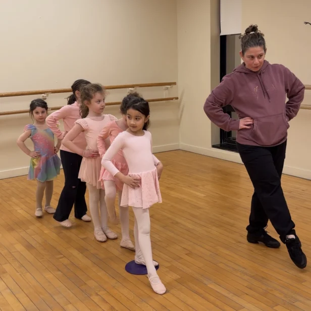 A ballet instructor demonstrates a pose while five young girls in dance attire line up behind her, mimicking the pose in a studio with wooden floors and mirrored walls.