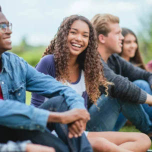 A group of five teenagers sit together on grass outdoors, smiling and talking, with greenery and sky in the background.