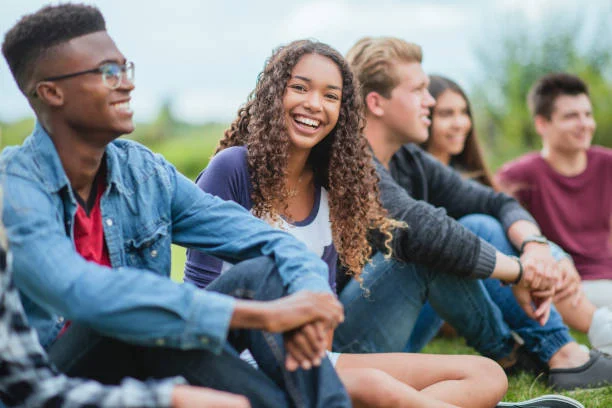 A group of five teenagers sit together on grass outdoors, smiling and talking, with greenery and sky in the background.