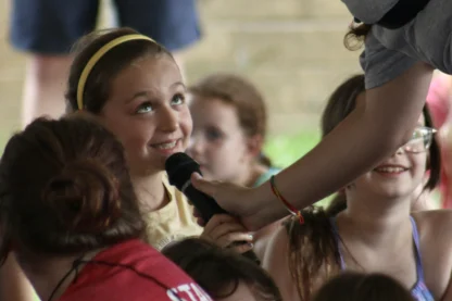A young girl smiles and looks up as she speaks into a microphone held by an adult during an outdoor event, with other children nearby.