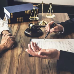 Two people sit at a desk with legal documents, a gavel, scales of justice, and law books, suggesting a legal consultation or discussion.