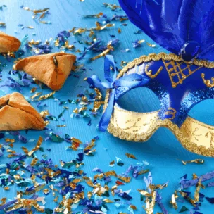 A blue and gold masquerade mask with feathers lies on a blue surface scattered with confetti, next to three triangular hamantaschen cookies.