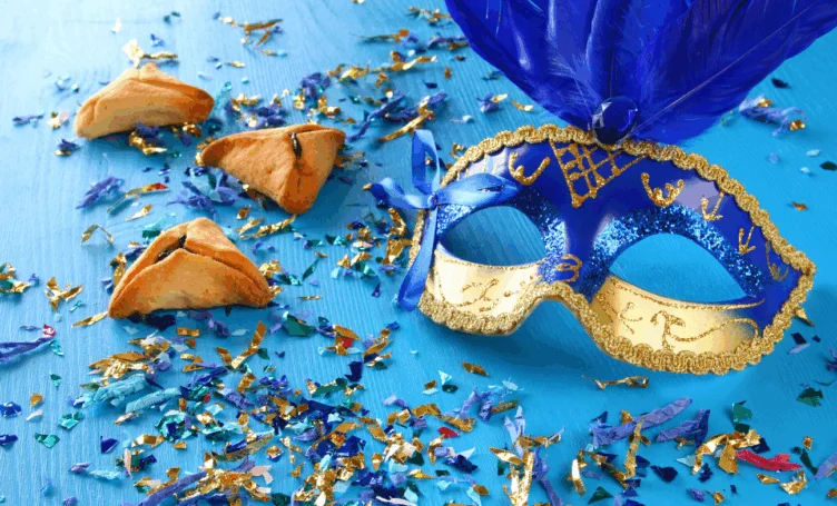 A blue and gold masquerade mask with feathers lies on a blue surface scattered with confetti, next to three triangular hamantaschen cookies.