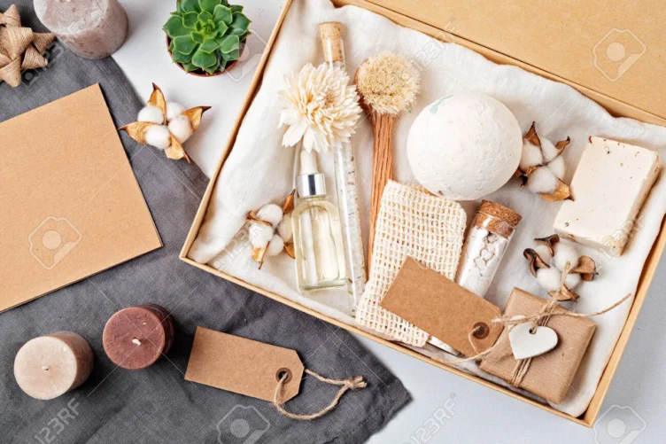 A gift box containing bath and spa items such as soap, a brush, a bath bomb, a washcloth, a bottle, candles, cotton flowers, and blank tags on a table.