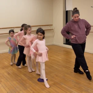 A ballet instructor demonstrates a pose while five young girls in dancewear line up and follow her lead in a studio.