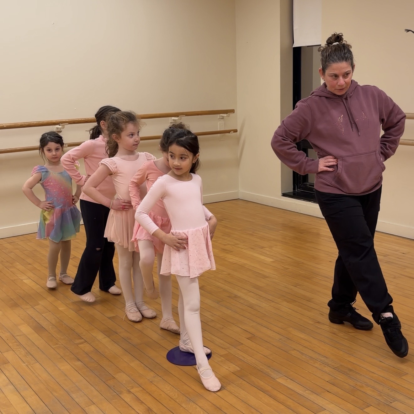 A ballet instructor demonstrates a pose while five young girls in dancewear line up and follow her lead in a studio.