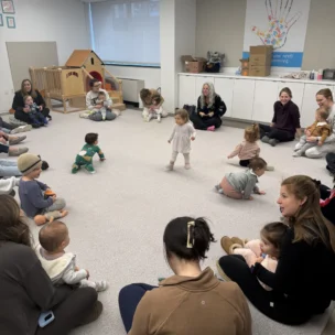 A group of adults and toddlers sit in a large circle on the floor of a daycare room while a toddler walks in the center.