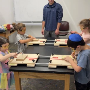 A group of young children work on woodworking projects at a table while an adult supervises in a classroom setting.