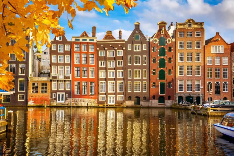 Row of traditional narrow Dutch buildings reflected in a canal, with autumn leaves in the foreground and a boat docked at the water's edge.