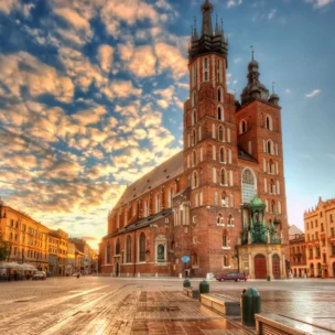 St. Mary's Basilica in Kraków, Poland, stands in a large town square at sunset with a partly cloudy sky and historic buildings surrounding the area.