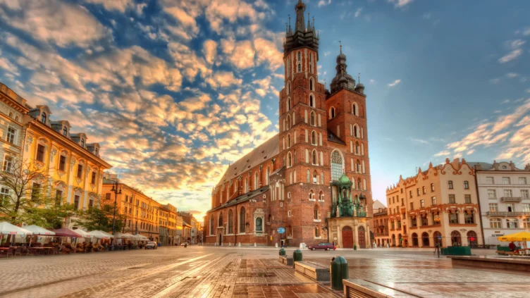 St. Mary's Basilica in Kraków, Poland, stands in a large town square at sunset with a partly cloudy sky and historic buildings surrounding the area.