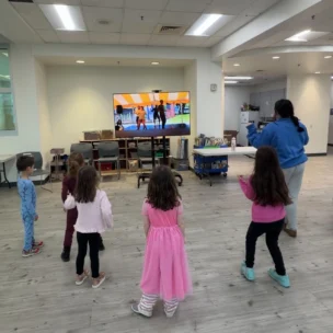 Five young children and one adult stand in a spacious room, following a dance routine displayed on a large TV screen at the front.