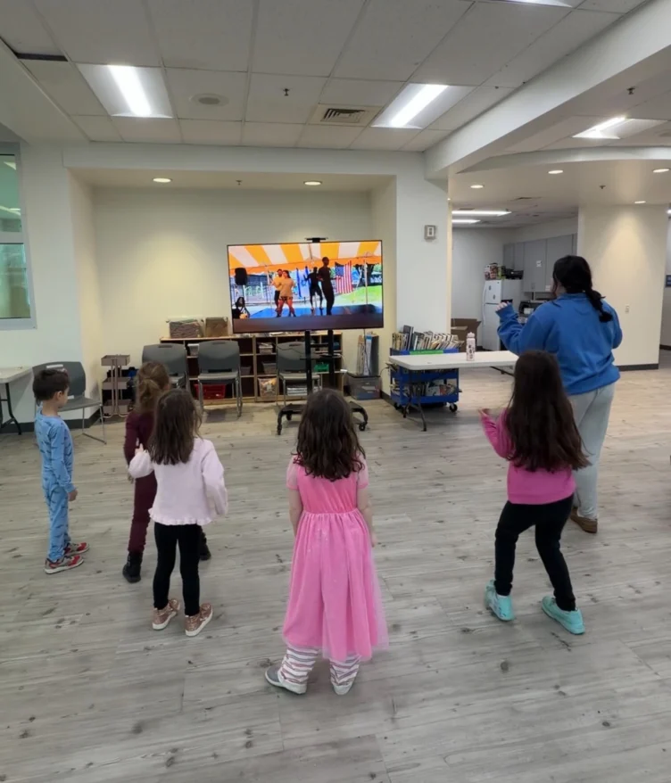 Five young children and one adult stand in a spacious room, following a dance routine displayed on a large TV screen at the front.