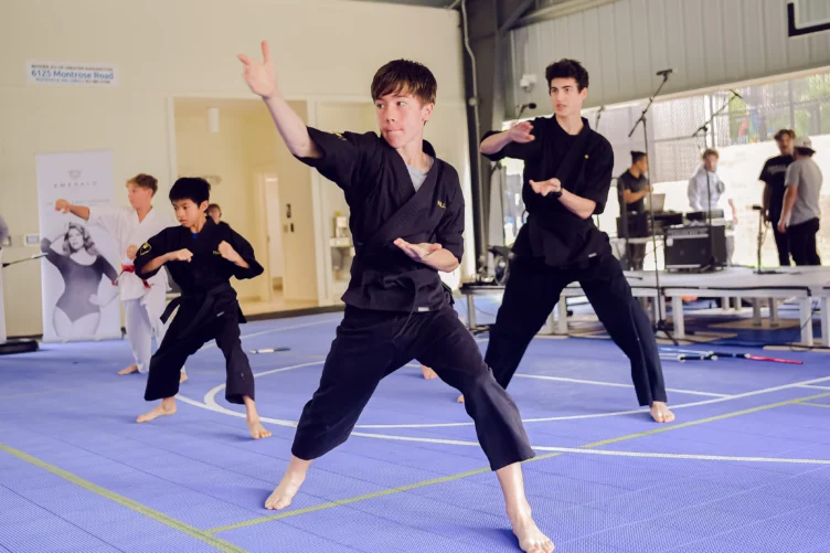 A group of martial arts students in black uniforms practice forms on a blue mat in an indoor gym.