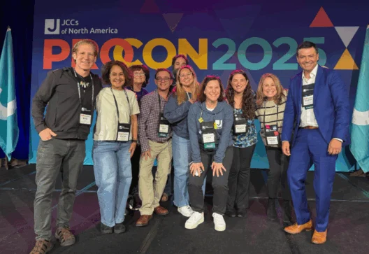 A group of ten people pose together on stage at the JCCs of North America ProCon 2025 event, standing in front of a colorful event backdrop.