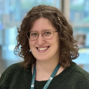 A person with curly brown hair and glasses, wearing a dark green sweater and a lanyard, smiles at the camera in an indoor setting.