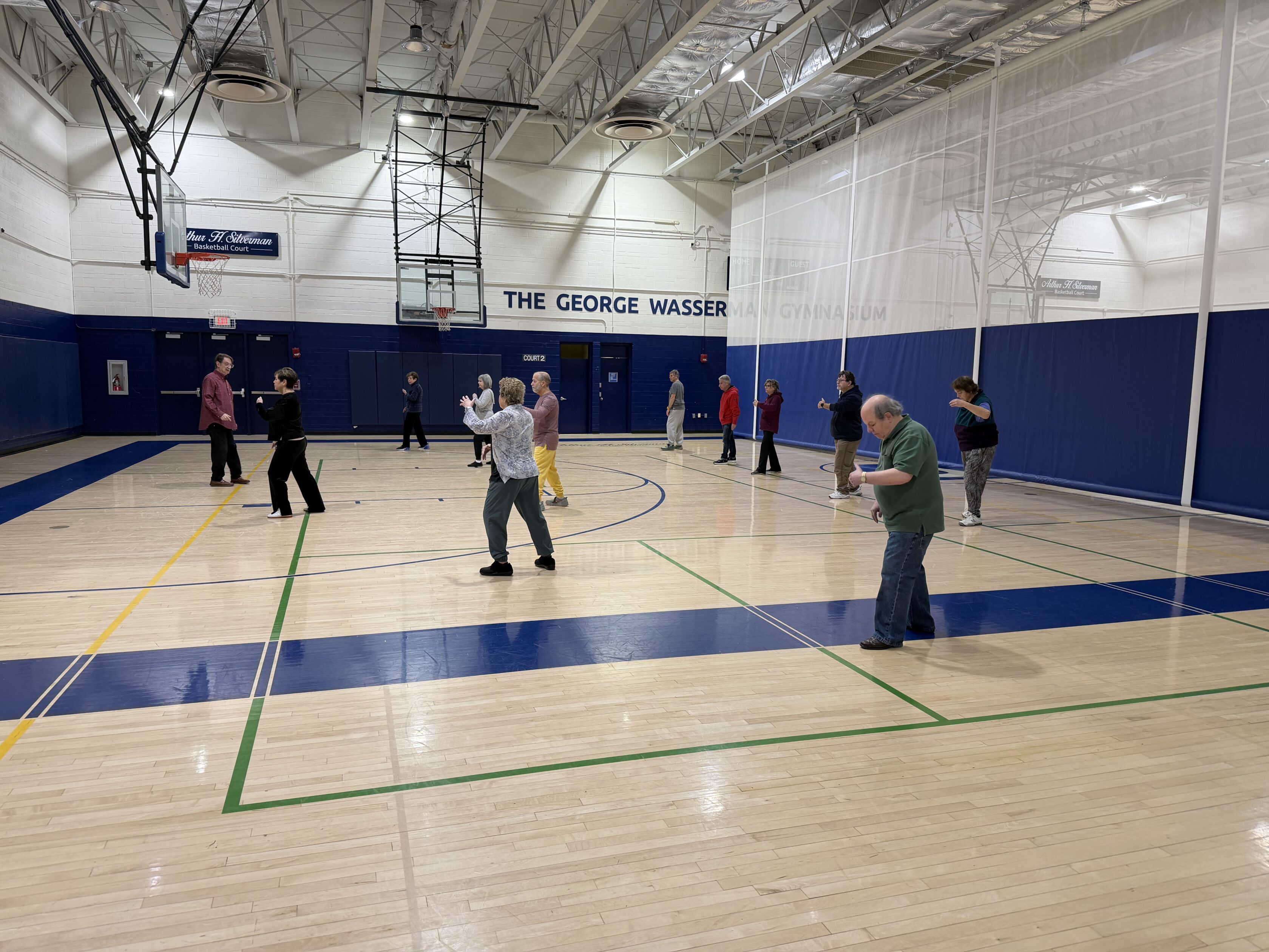 A group of people practice tai chi in a gymnasium, spaced out across the basketball court under bright overhead lights.