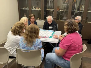 Seven adults sit around a table playing a board game in a room with display cabinets in the background.