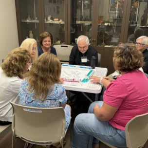 Seven adults sit around a table playing a board game in a room with display cabinets in the background.