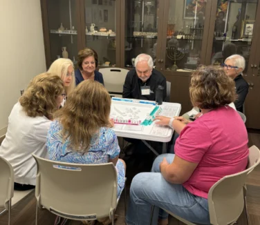 Seven adults sit around a table playing a board game in a room with display cabinets in the background.