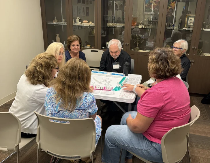 Seven adults sit around a table playing a board game in a room with display cabinets in the background.