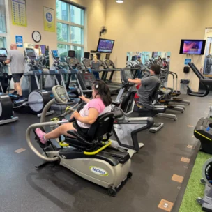 People exercise on various cardio machines in a gym, including treadmills, ellipticals, and recumbent steppers; TVs are mounted on the wall, and sunlight enters through large windows.