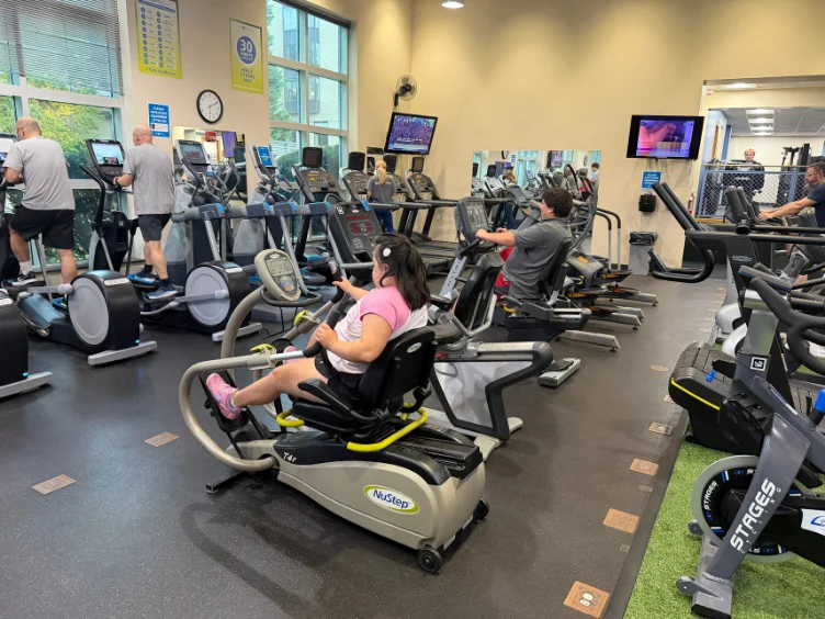 People exercise on various cardio machines in a gym, including treadmills, ellipticals, and recumbent steppers; TVs are mounted on the wall, and sunlight enters through large windows.