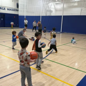 Young children and adults participate in a basketball activity inside a gymnasium, with some kids holding basketballs and others moving around the court.
