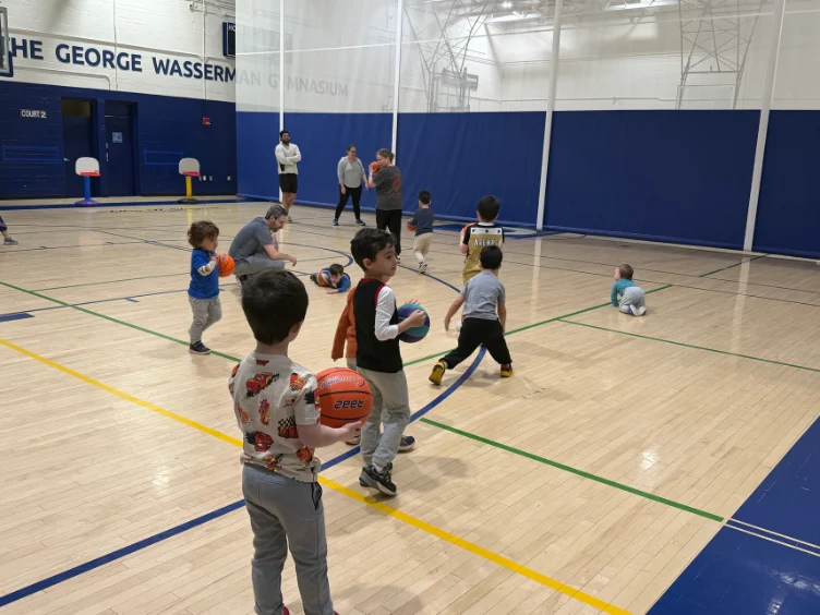 Young children and adults participate in a basketball activity inside a gymnasium, with some kids holding basketballs and others moving around the court.