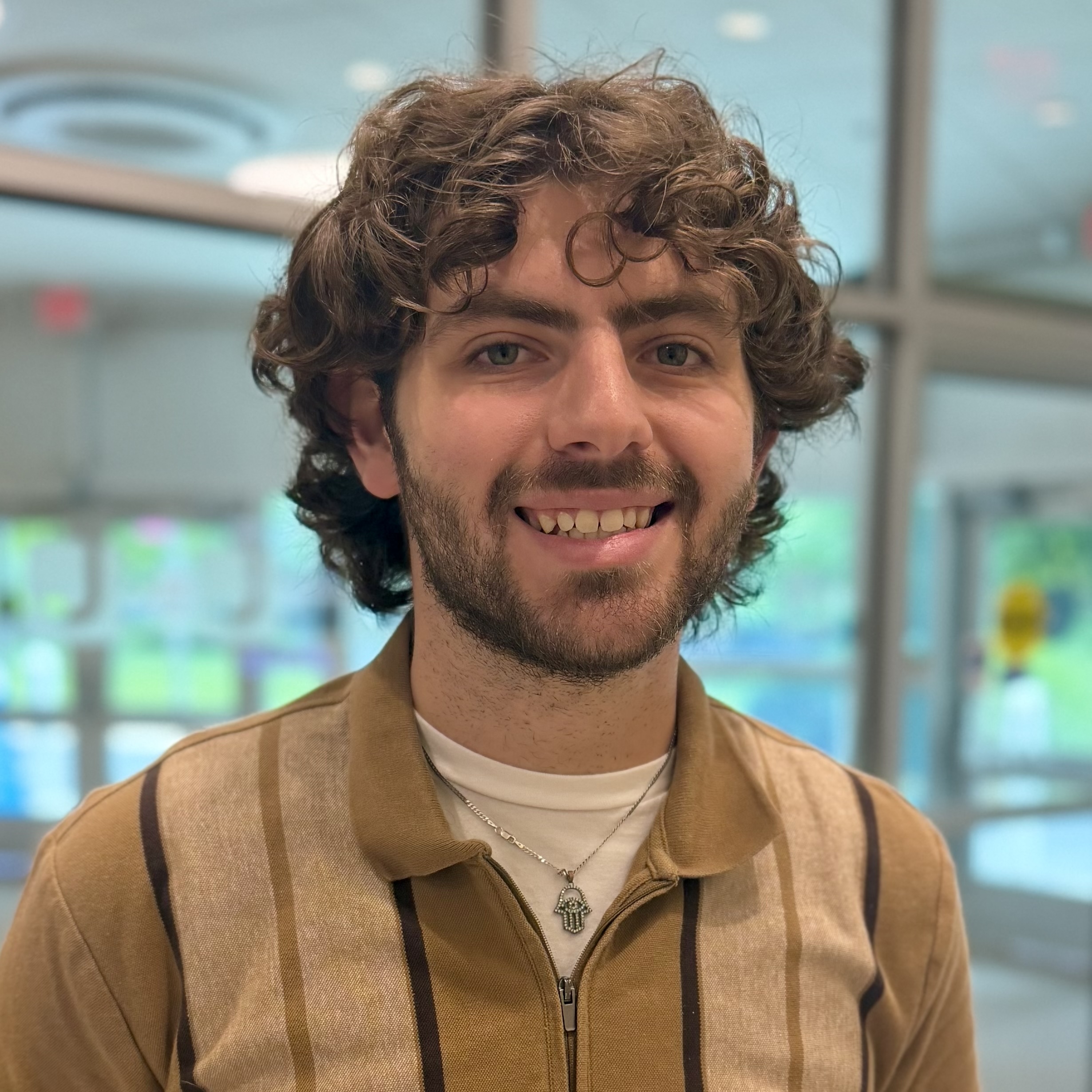 A person with curly brown hair and a beard smiles indoors, wearing a brown collared shirt and a necklace with a hand-shaped pendant. Glass doors and greenery are visible in the background.