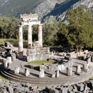 Ruins of a circular ancient Greek temple with columns and stone remains, surrounded by trees and hills in the background.