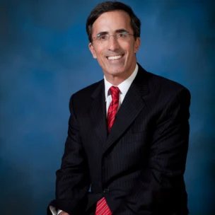 Man in a dark pinstripe suit with a red striped tie sitting and smiling against a blue background.
