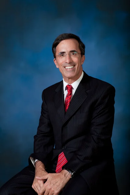 Man in a dark pinstripe suit with a red striped tie sitting and smiling against a blue background.