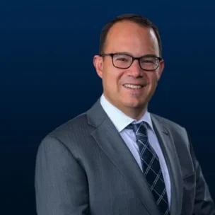 A man in a gray suit, white shirt, and patterned tie stands in front of a dark blue background, smiling at the camera.