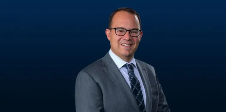 A man in a gray suit, white shirt, and patterned tie stands in front of a dark blue background, smiling at the camera.