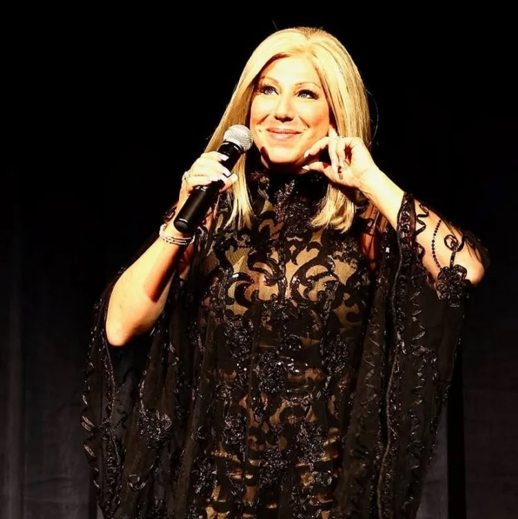 A person with long blonde hair holds a microphone and smiles on stage, wearing a black lace dress with intricate patterns, against a dark background.