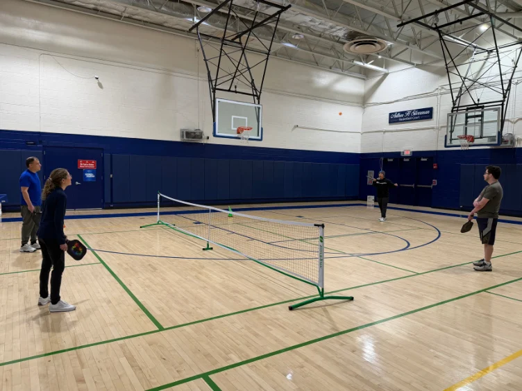 Four people play pickleball in a gymnasium. Two players are on each side of a portable net on a basketball court. Basketball hoops are visible above.